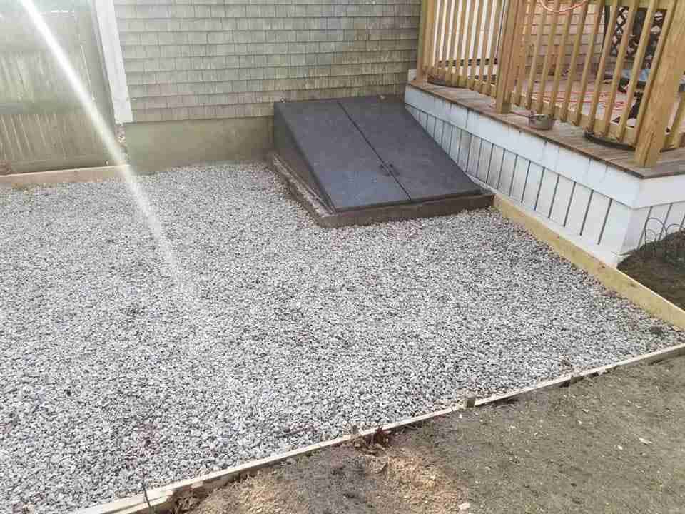 Gravel yard with a small black wooden ramp beside a brick house and deck steps.
