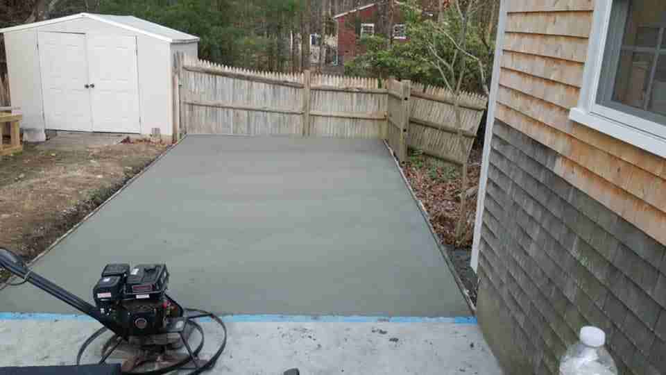 Freshly poured concrete patio beside a house, with a fence, shed, and a power trowel in the foreground.