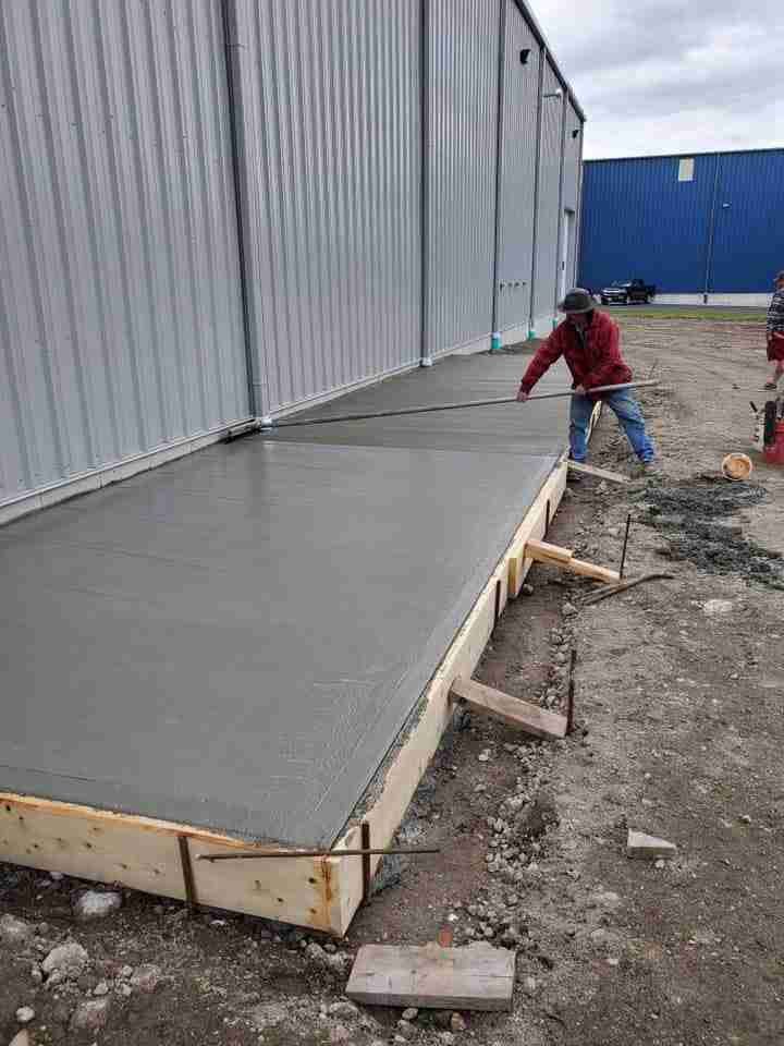 Worker smoothing wet concrete ramp beside a metal building at a construction site