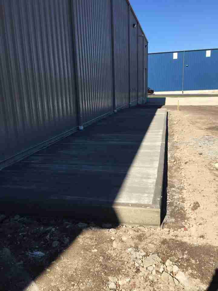 Concrete ramp beside a dark corrugated metal building on a gravel lot under bright sunlight