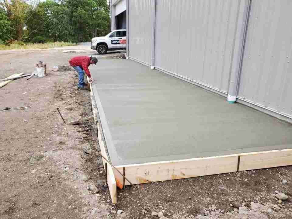 Worker smoothing fresh concrete slab beside a metal building