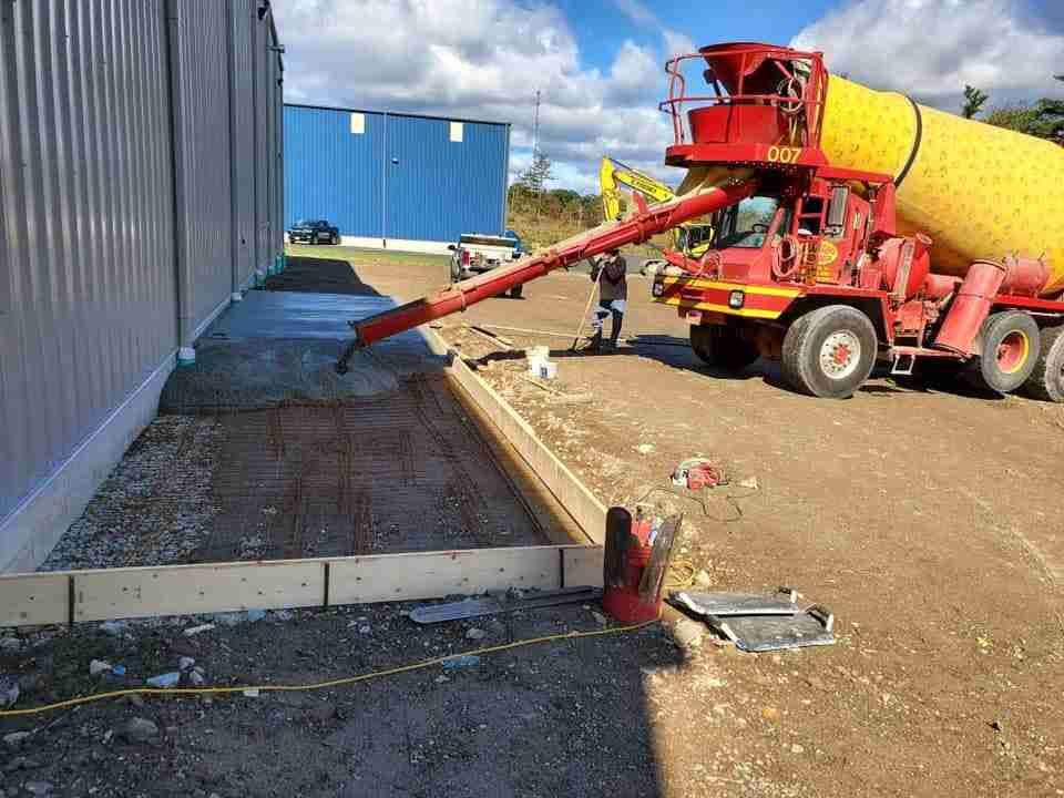 Concrete being poured from a red mixer truck into a trench beside a corrugated metal building.