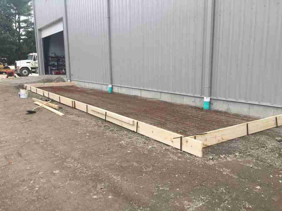 Wooden-framed concrete slab foundation beside a gray metal building under construction.