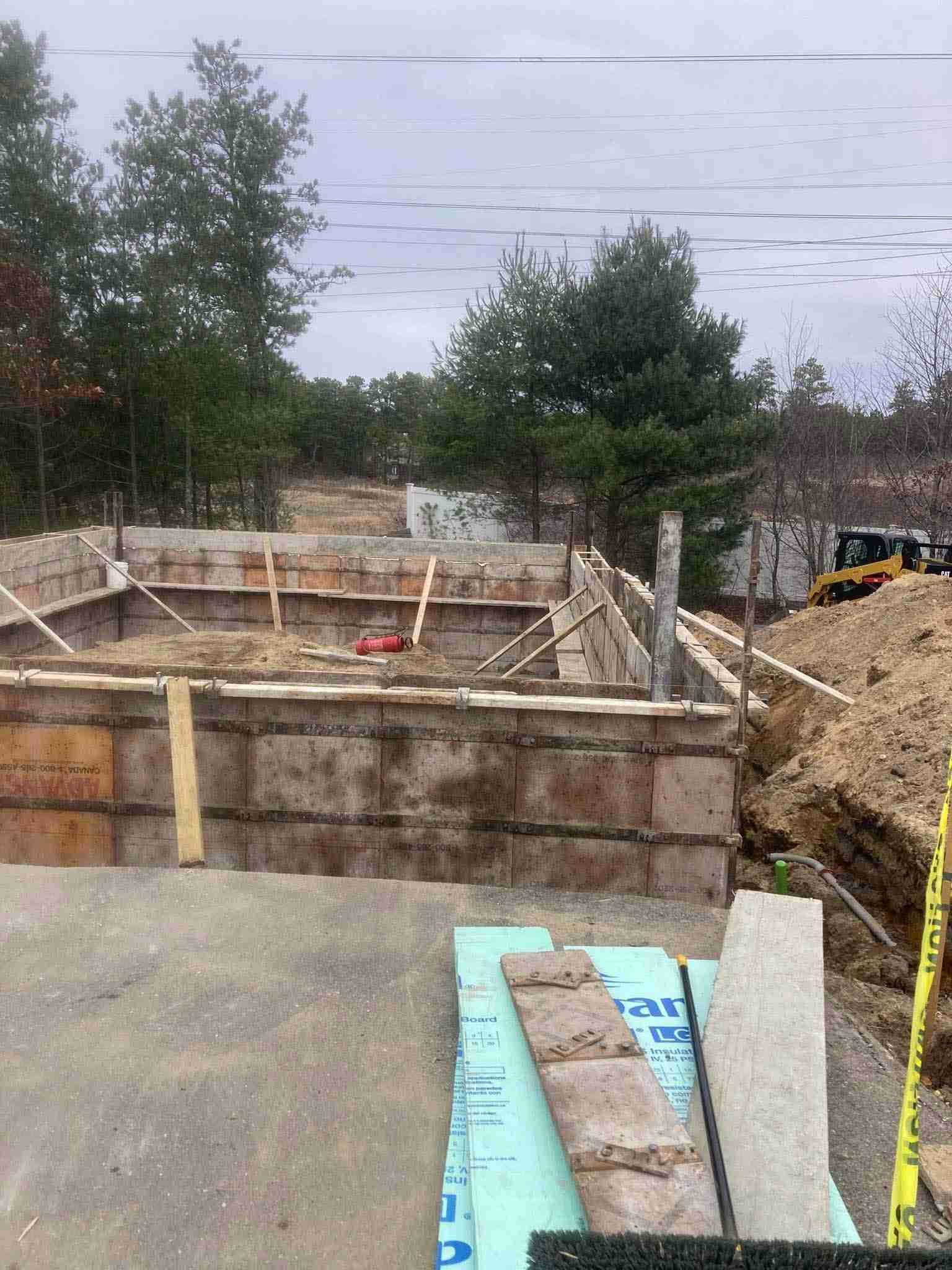 Construction site with a concrete foundation, wooden formwork, dirt piles, and trees under a cloudy sky