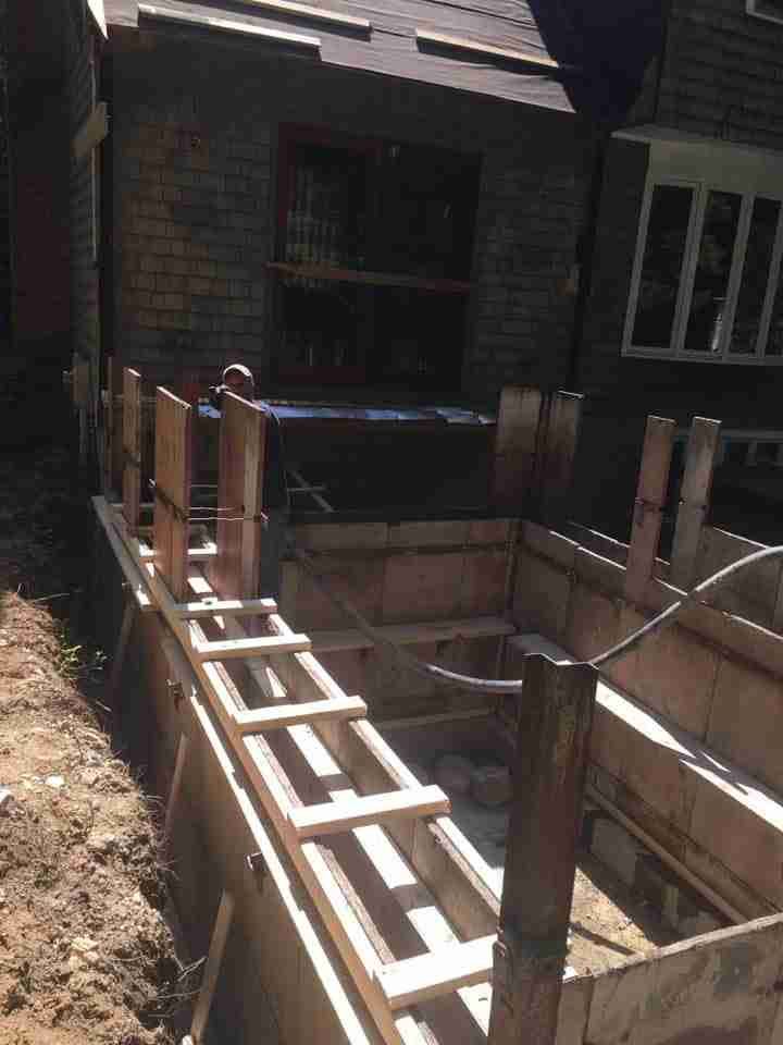 Brick house construction with a narrow concrete walkway and wooden railing framework alongside a trench