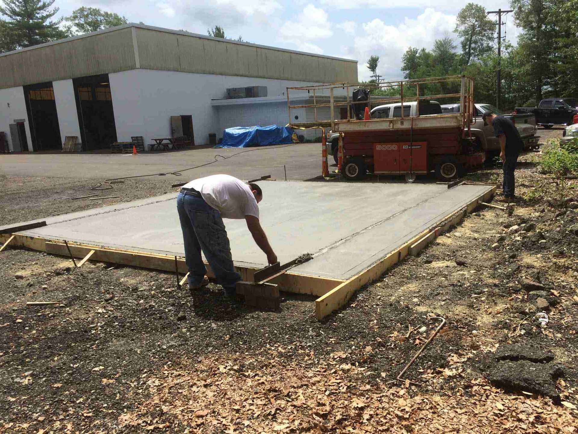 Worker leveling freshly poured concrete slab at a construction site with a red truck nearby