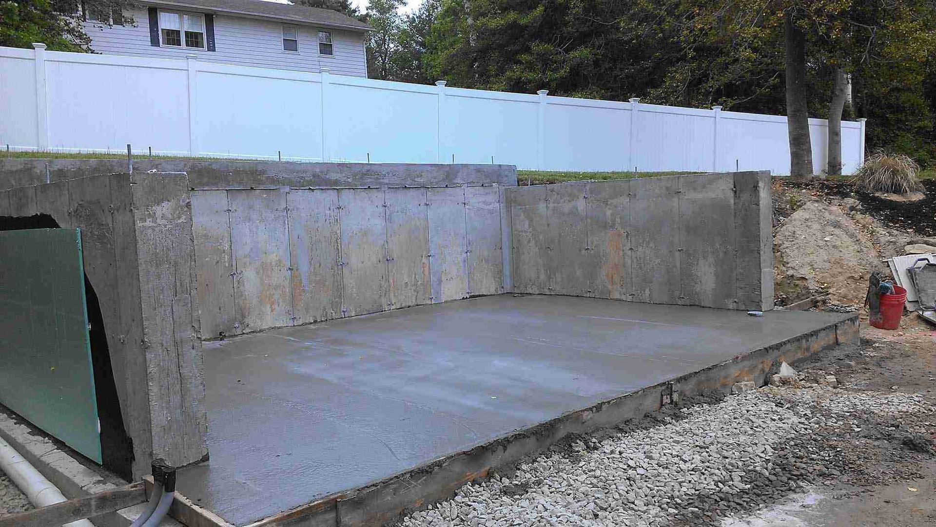 Concrete foundation slab and retaining walls under construction beside a white fence.