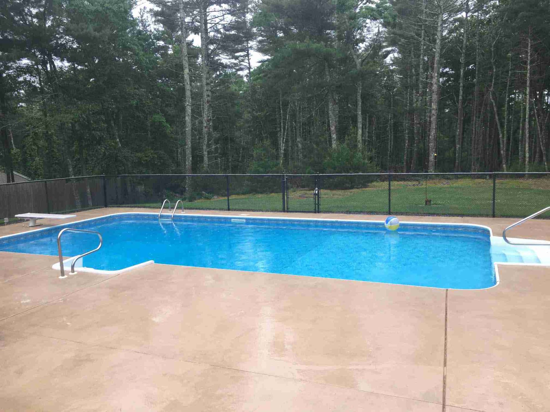 Backyard swimming pool with blue water, beige deck, and trees beyond a black fence