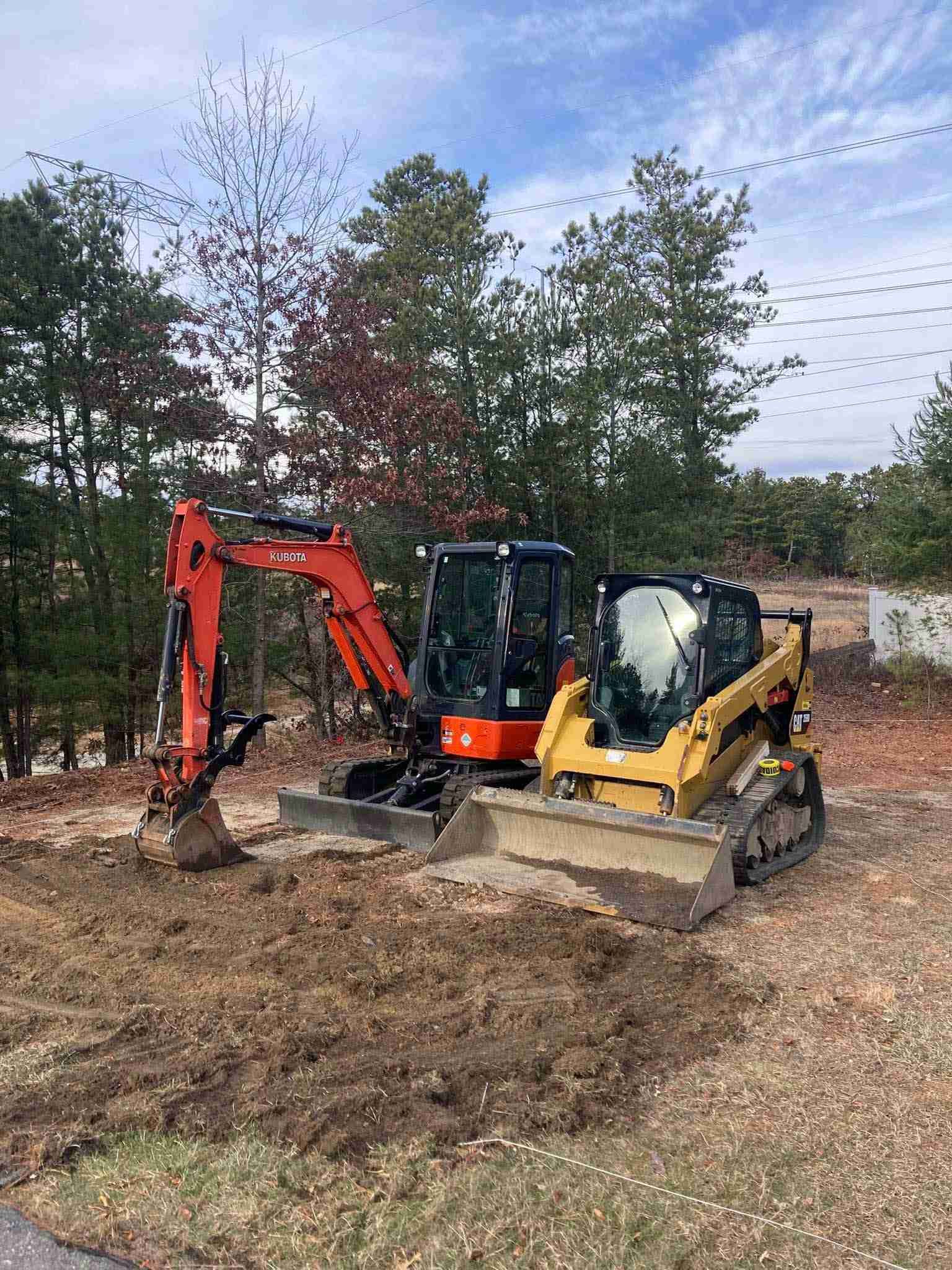 Yellow and orange mini excavators parked on a muddy construction site with trees in the background