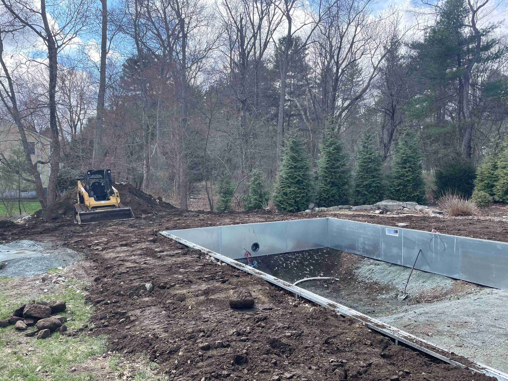 Backyard pool construction with a concrete basin, gravel, and a small excavator beside trees