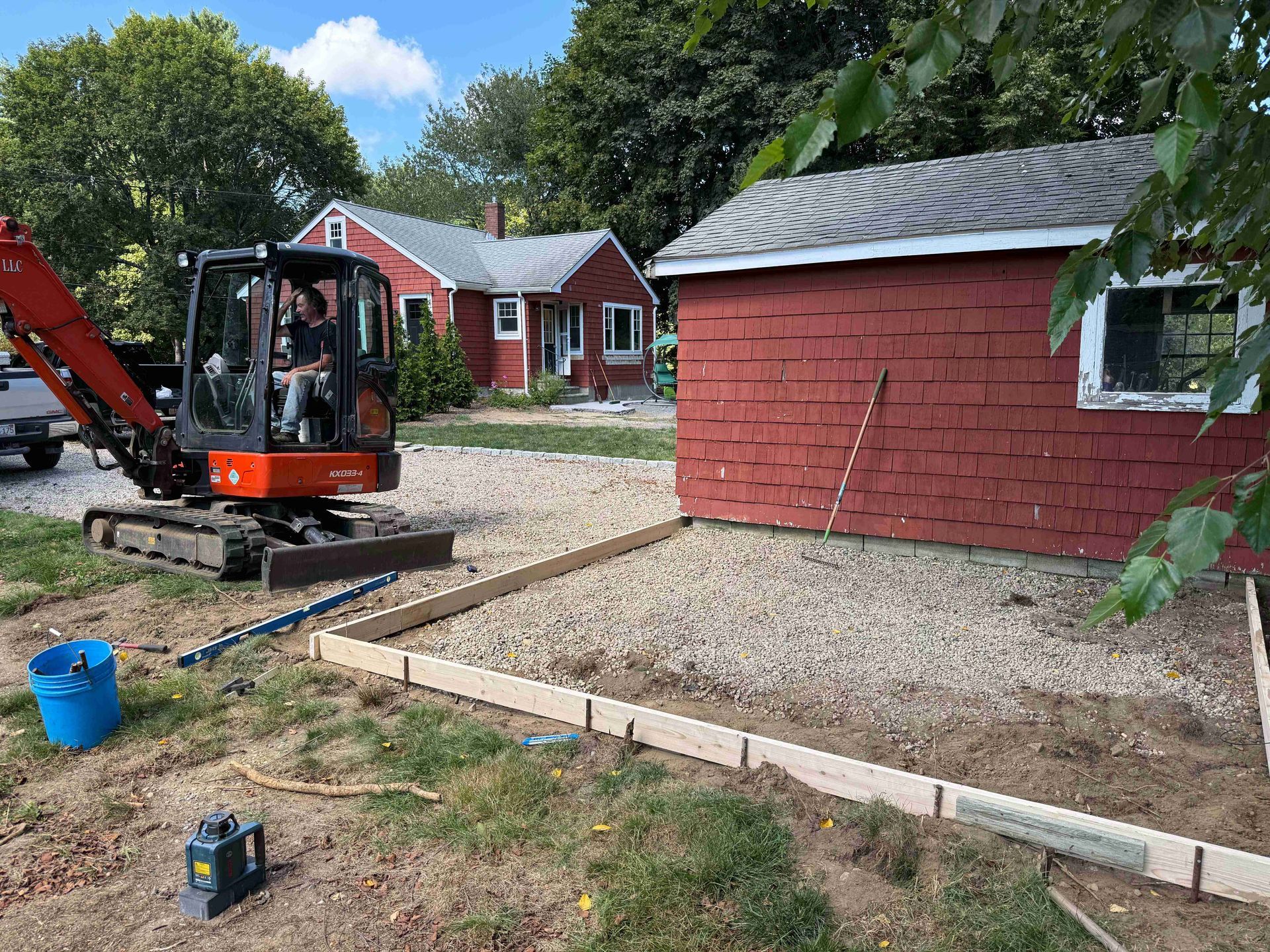 Mini excavator working beside a red shed in a gravel yard near a house and trees