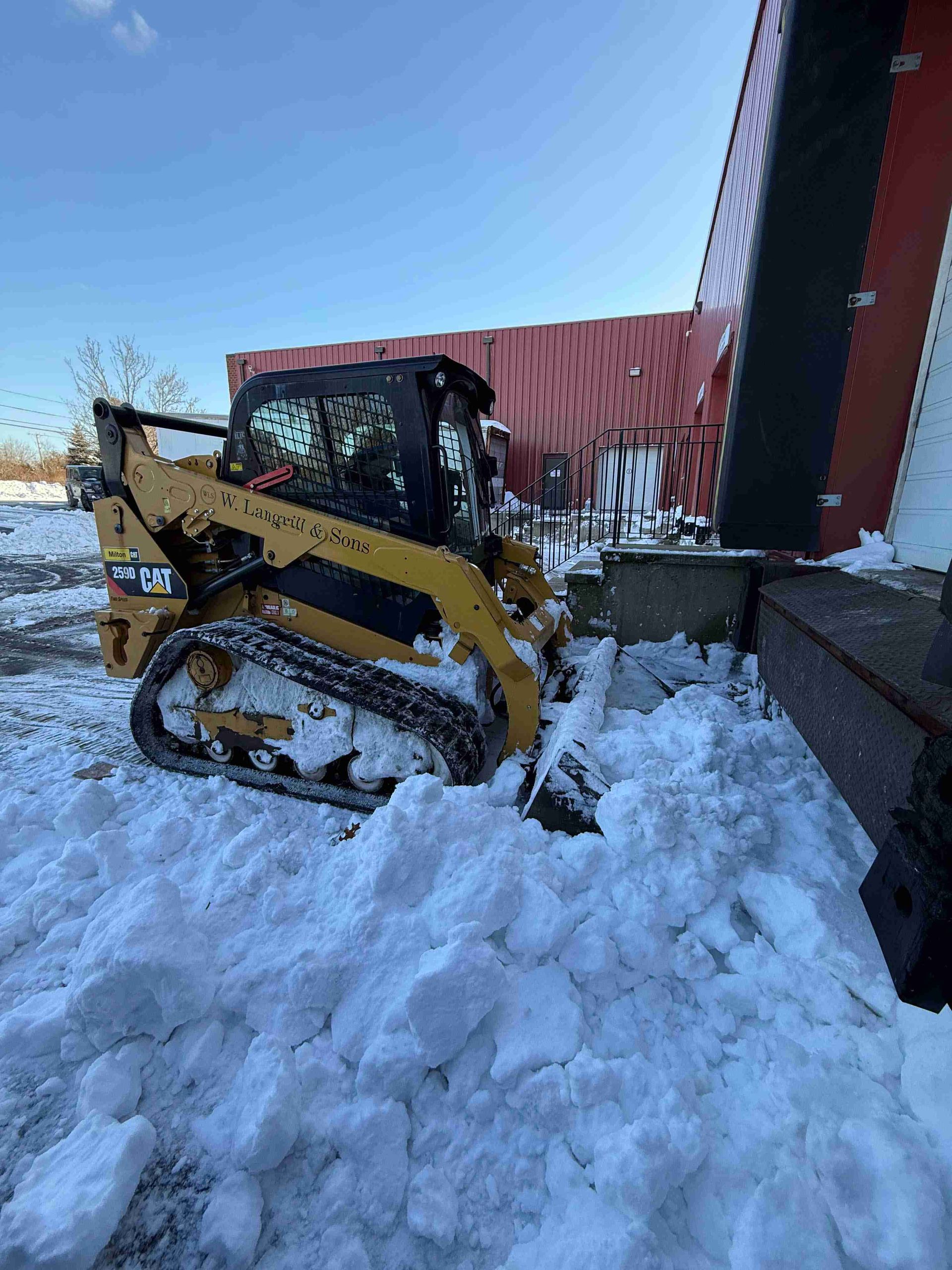 Yellow skid steer pushing through deep snow beside a red building on a snowy day