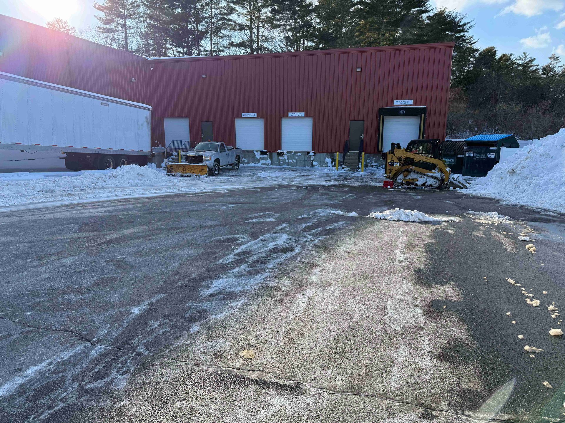 Snowy industrial yard with a red warehouse, parked equipment, and icy pavement in winter sunlight