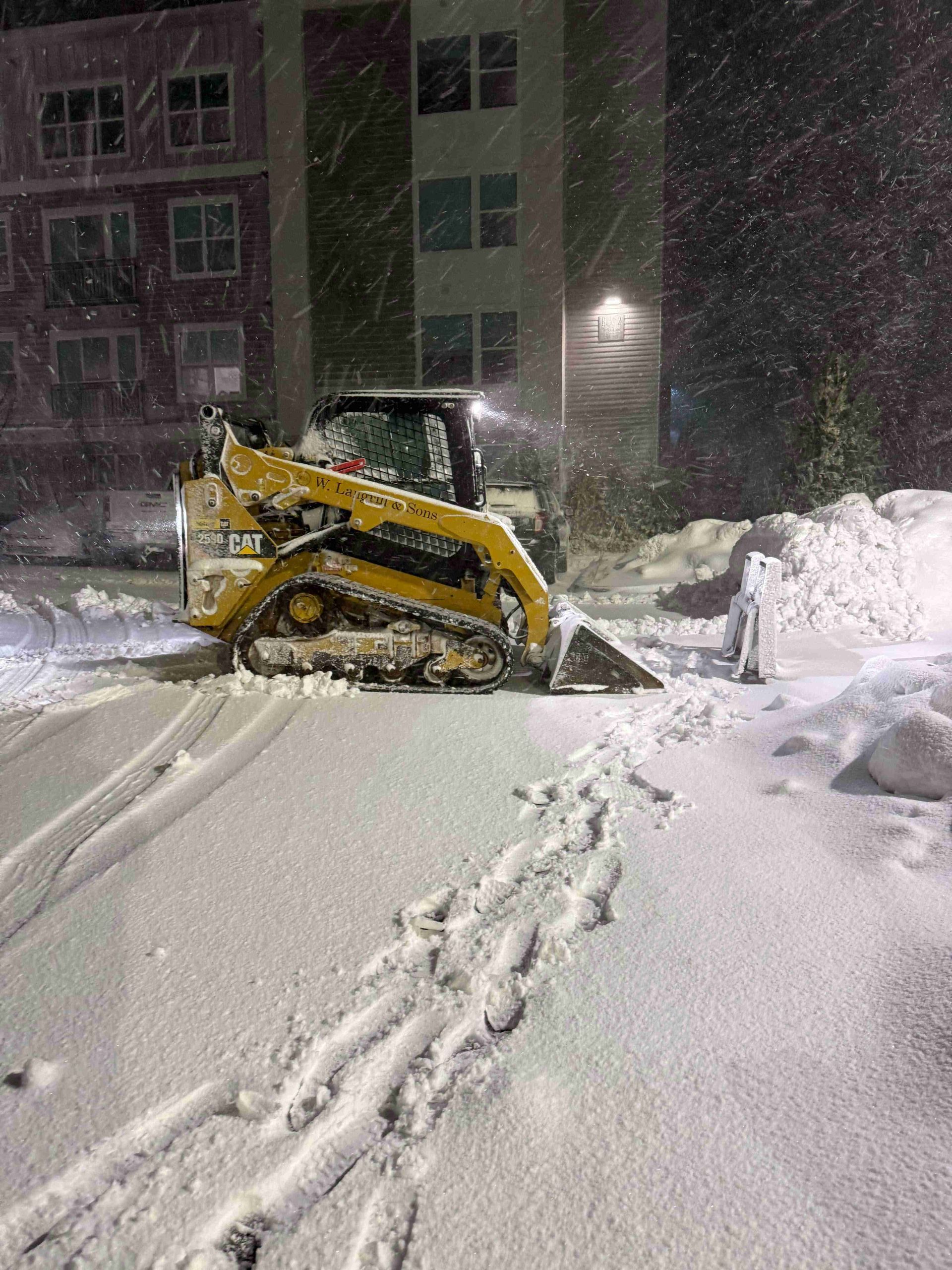 Yellow snowplow clearing a snowy apartment parking lot at night