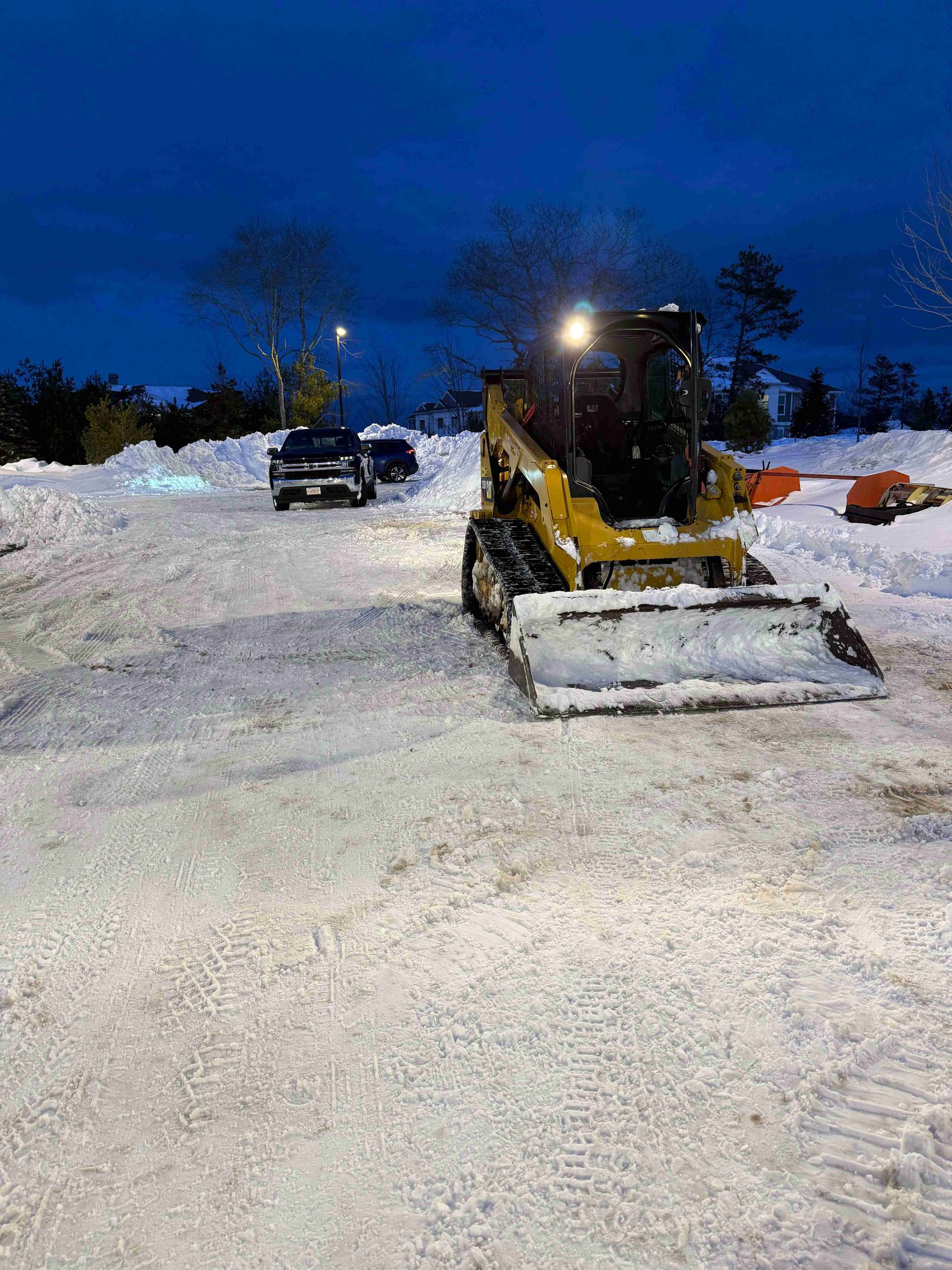 Yellow snowplow clearing a snowy lot at dusk, with parked vehicles and trees in the background