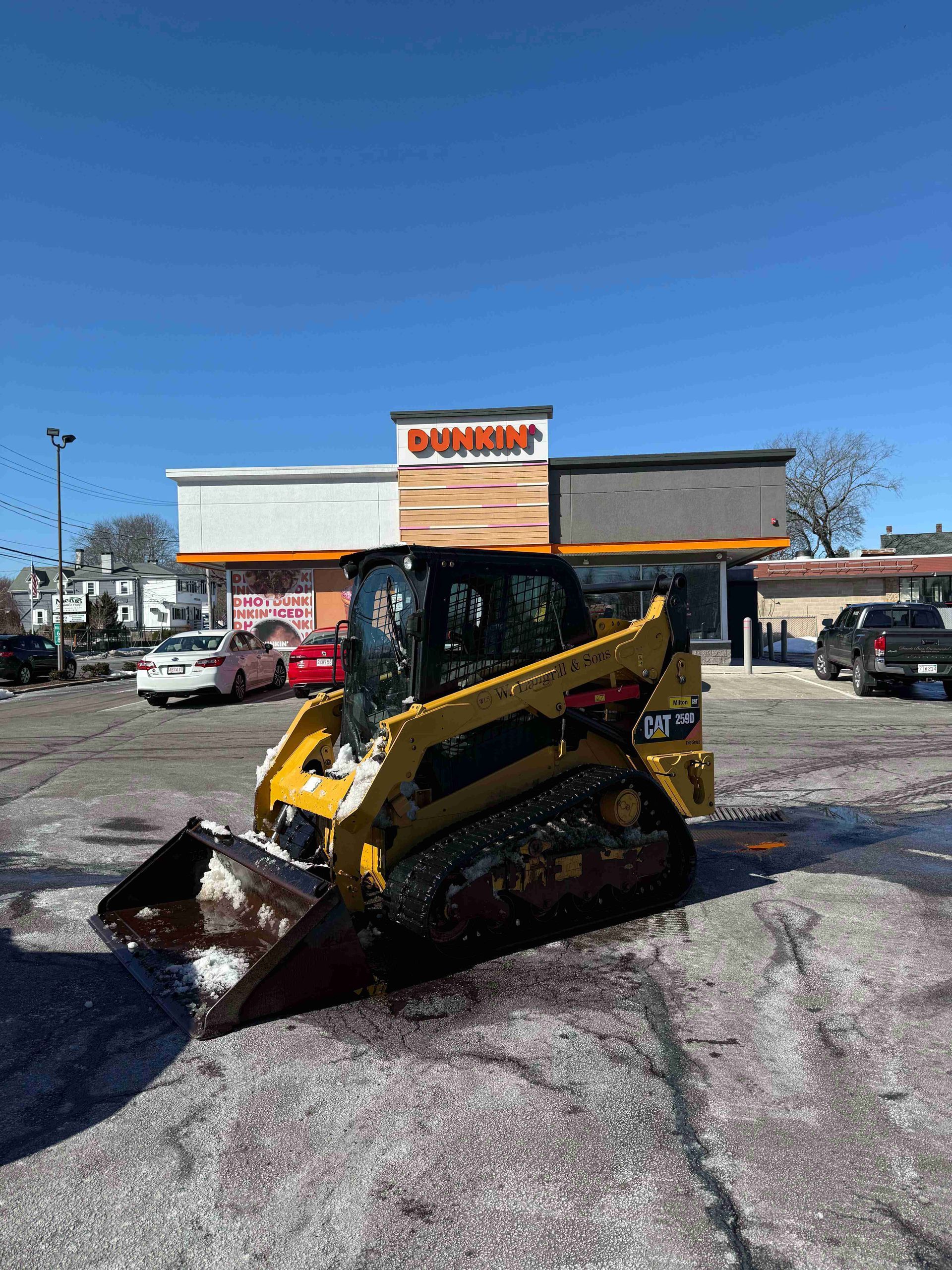 Yellow skid steer with front bucket parked in a snowy lot outside a store under a clear blue sky