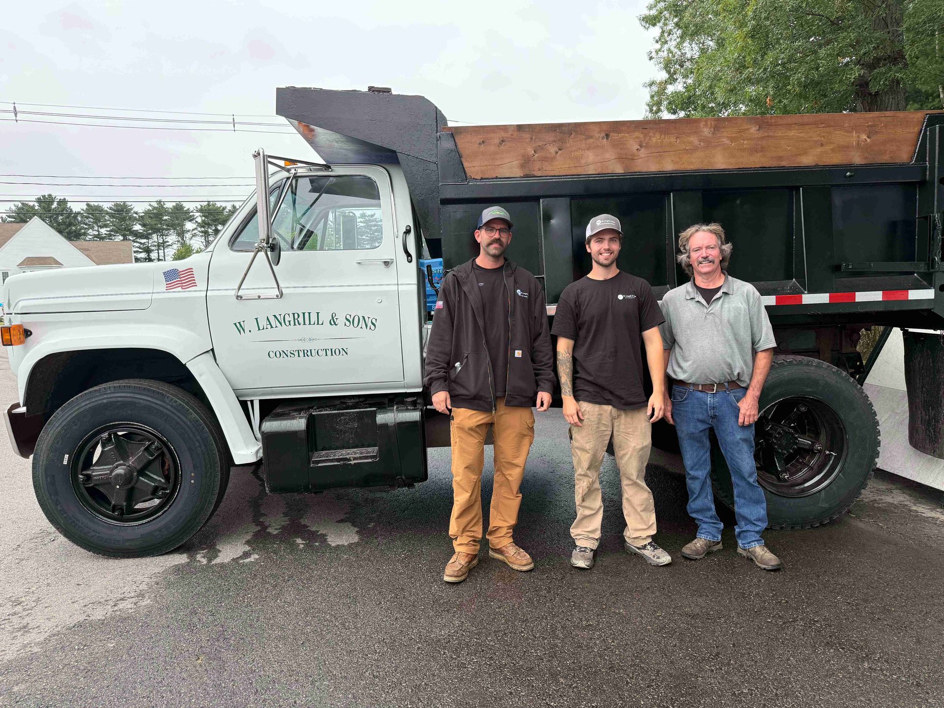 Three men stand in front of a white dump truck with a black bed on a wet road.