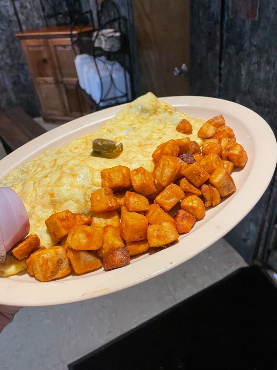 Plate of orange diced food with creamy mashed potatoes and a pickle, held outdoors