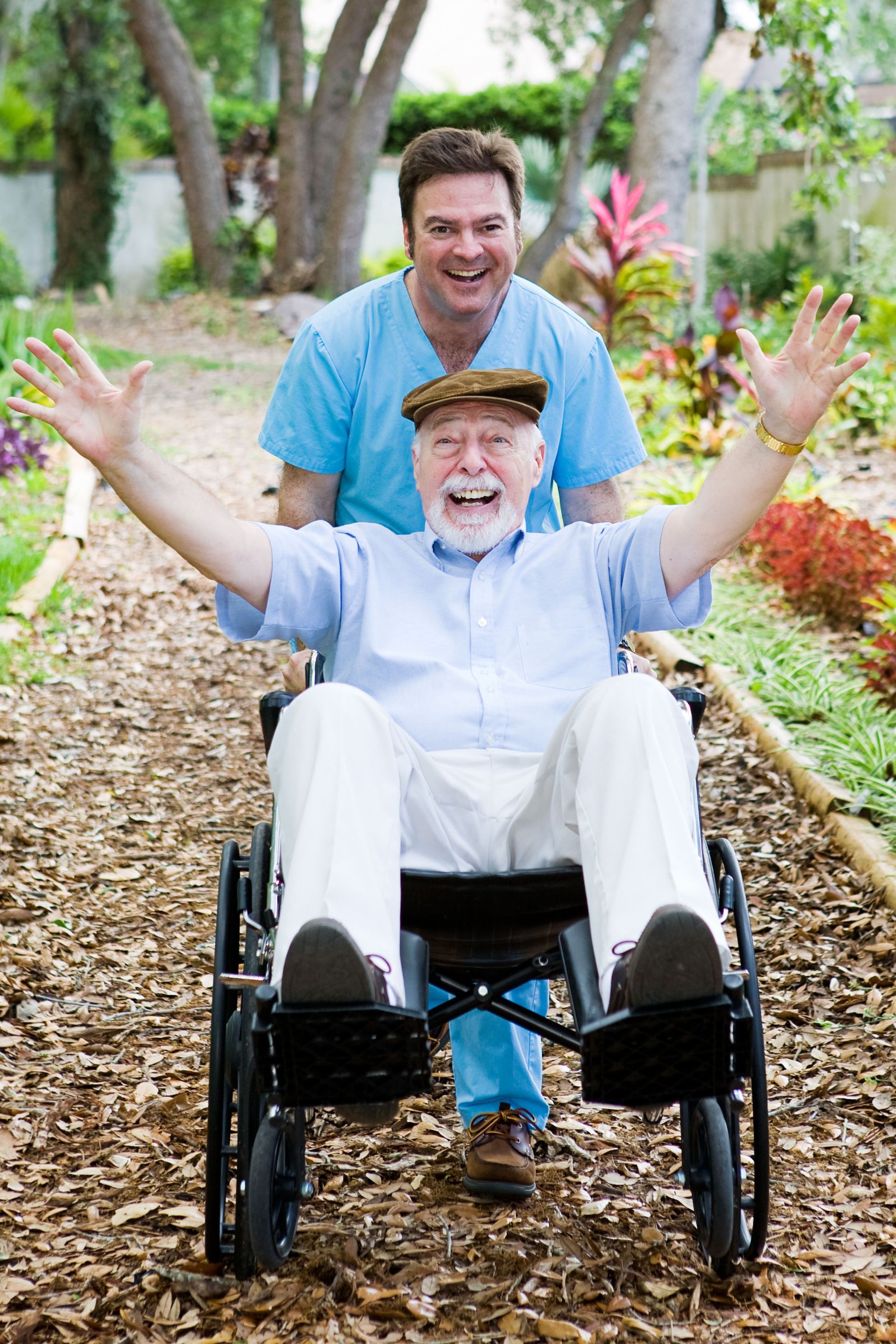 Older man in wheelchair with raised arms, smiling, pushed by another person on a garden path