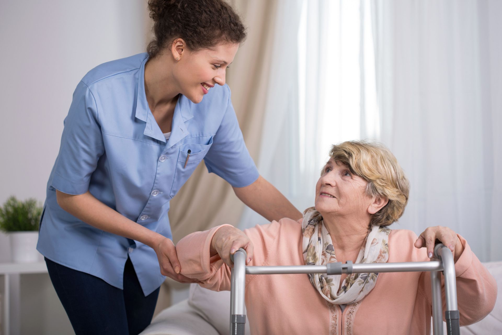 Caregiver helping an elderly woman walk with a walker in a bright room