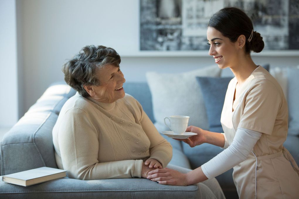 Caregiver serves tea to an elderly woman resting on a couch in a bright living room