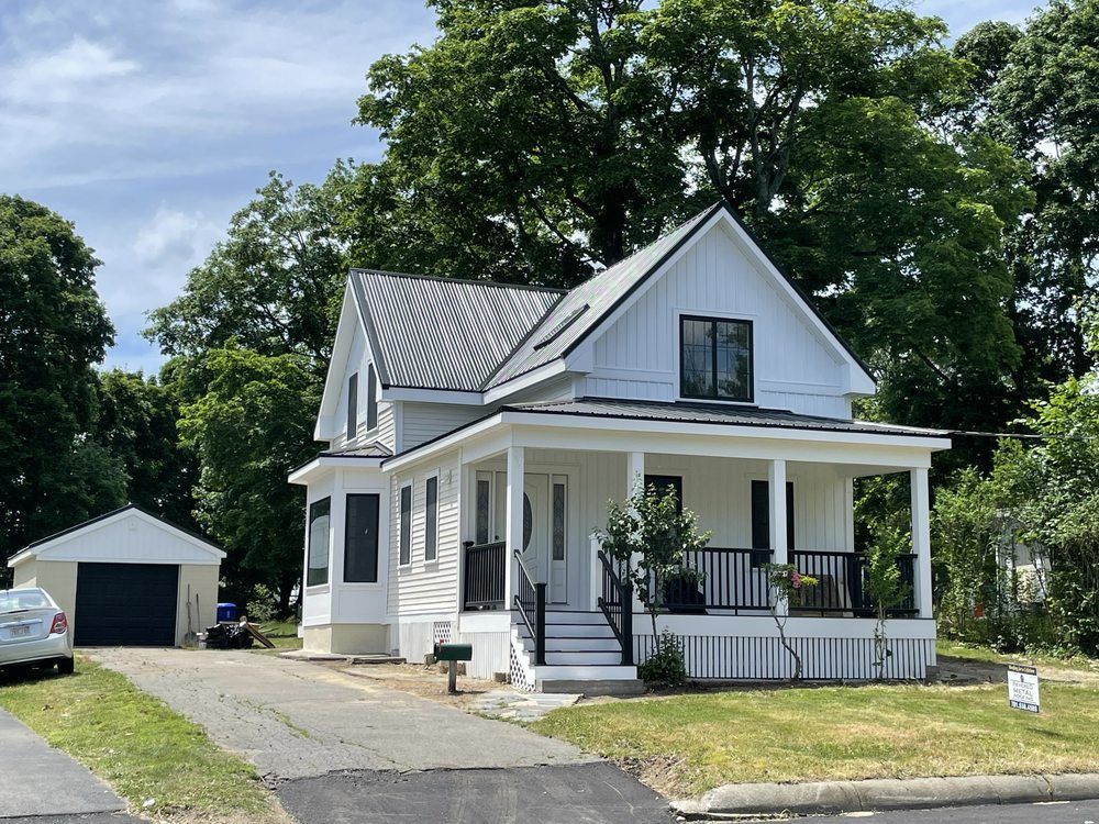 Workers on a black metal roof, installing components. Daytime, rural setting with trees and a dirt road visible.
