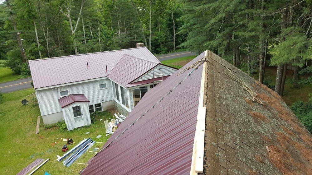 House with a new burgundy metal roof and an old, weathered roof section. Surrounded by trees.