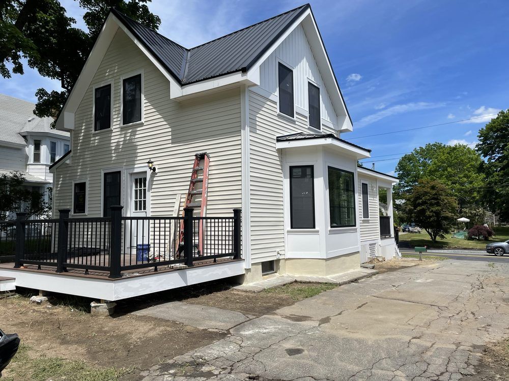Two-story house with black roof, black window frames, and beige siding; a deck with a black railing is in front.