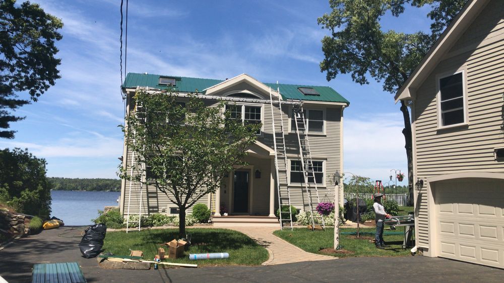 Two-story house with green roof next to a lake, with a ladder leaned on the side of the house.