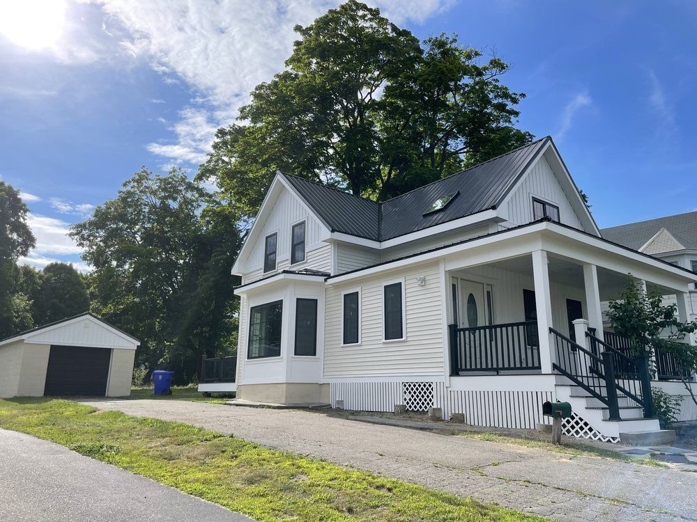 White house with black trim and roof, porch, garage, and driveway on a sunny day.