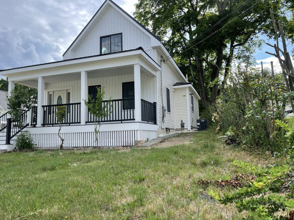 White farmhouse with black railings and porch on a grassy hill under a cloudy sky.