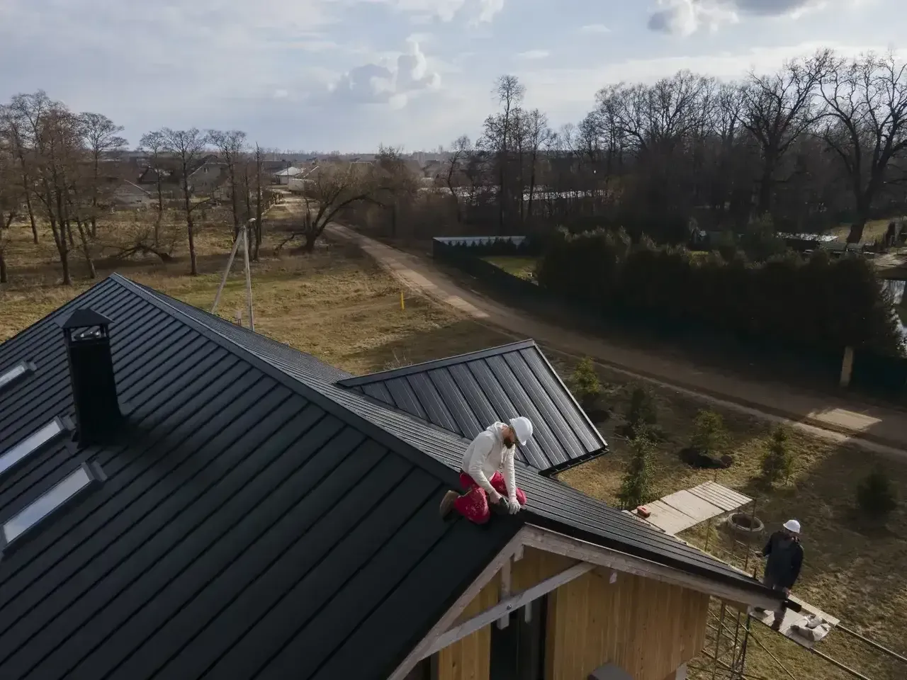 Workers on a black metal roof, installing components. Daytime, rural setting with trees and a dirt road visible.