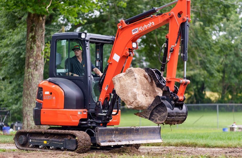 A man is driving a small excavator with a large rock in its bucket.