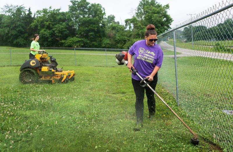 A woman is using a lawn mower to cut the grass next to a chain link fence.