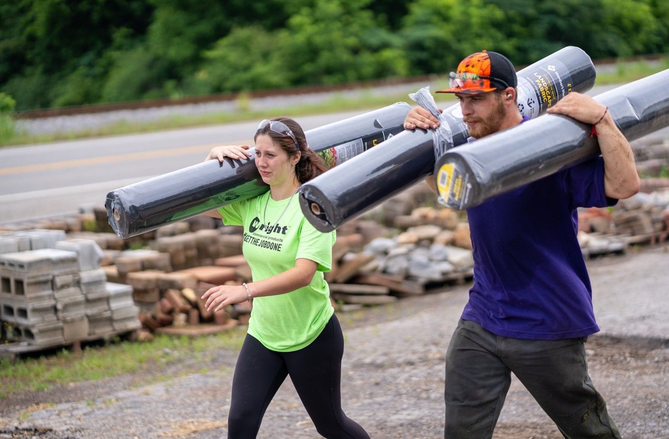 A man and a woman are carrying rolls of carpet on their shoulders.