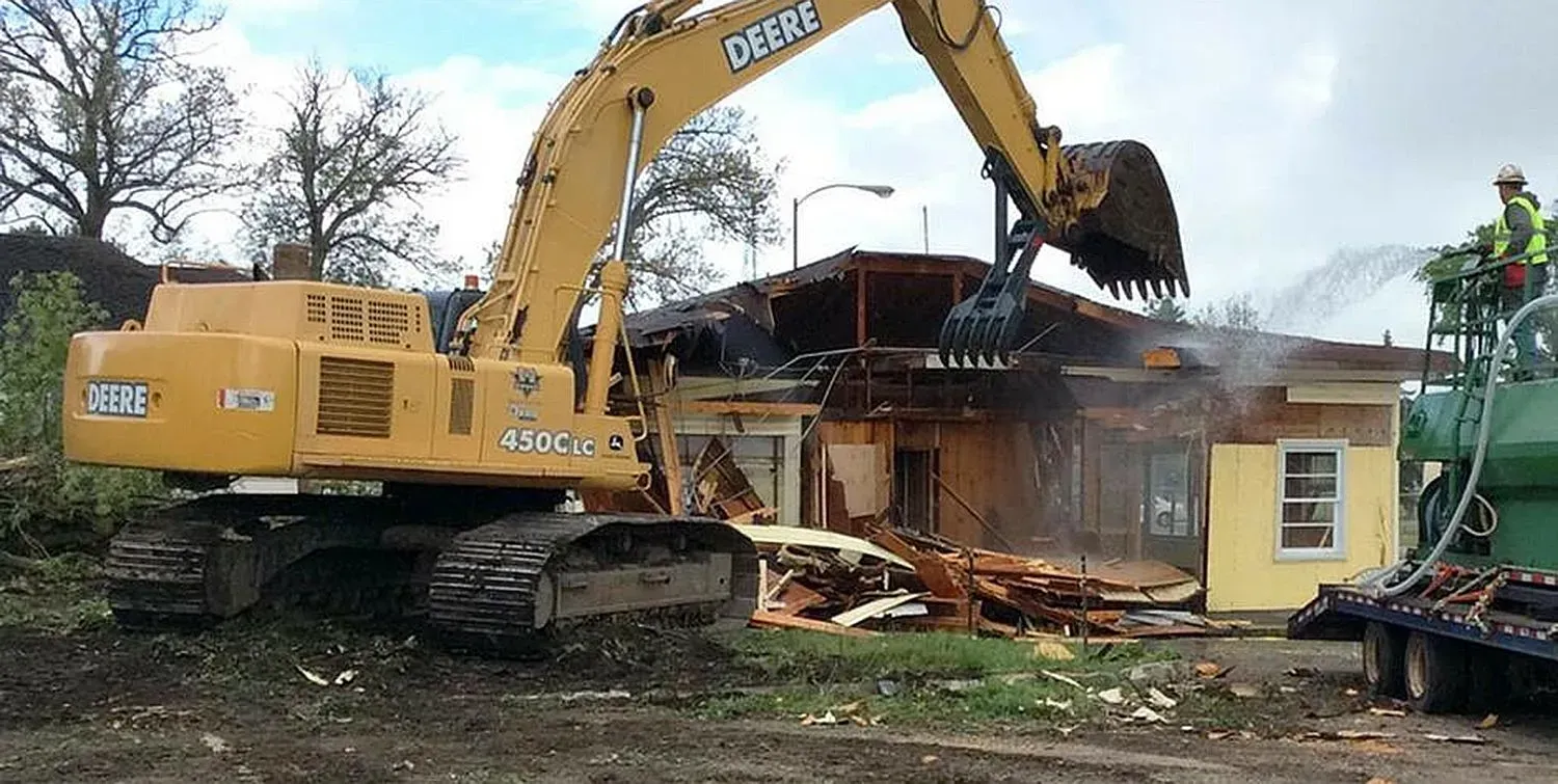 Yellow excavator demolishing a small house, with debris and dust at the construction site