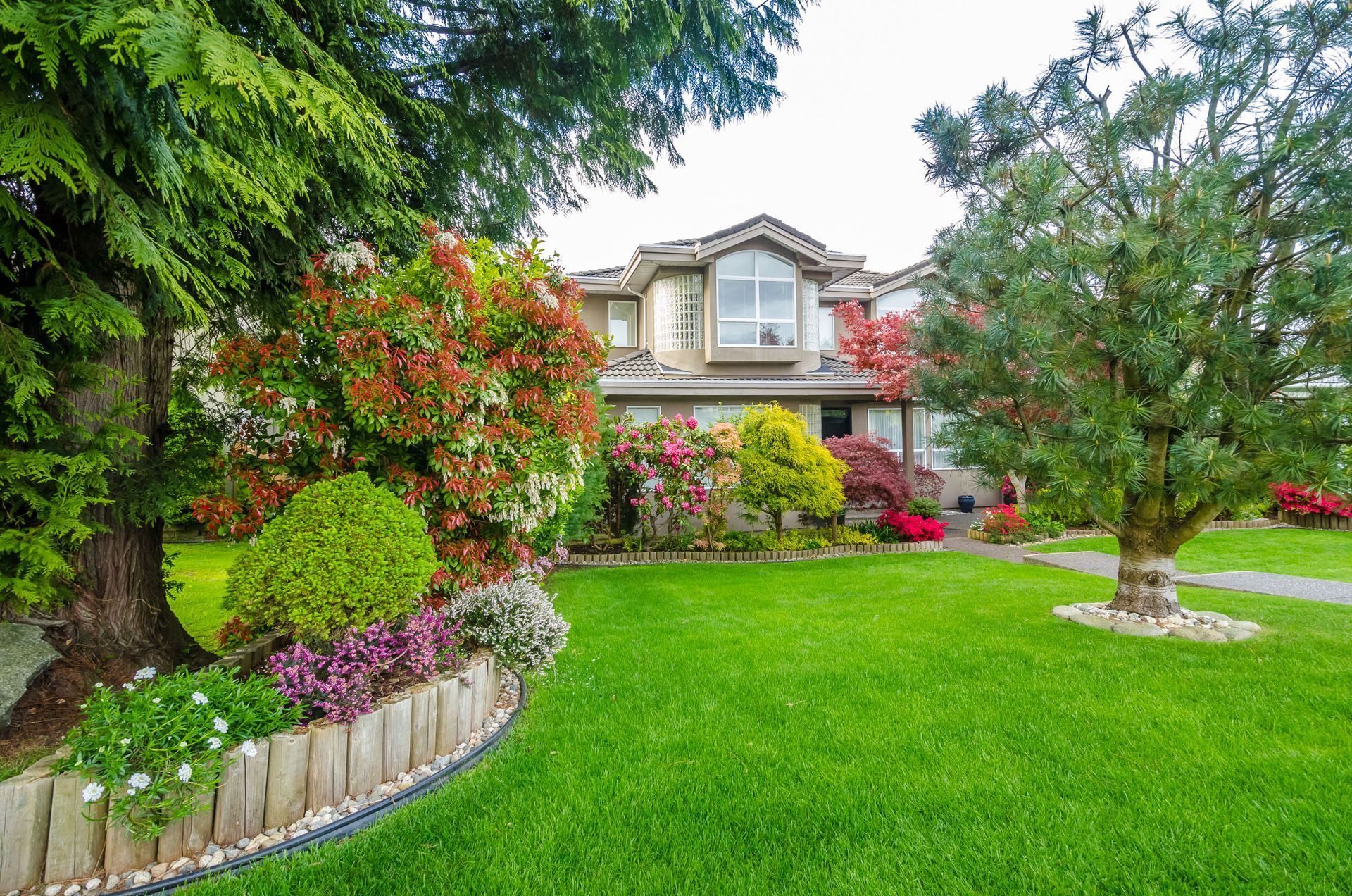 Lush green lawn with flowering bushes and trees in front of a two-story house with a decorative arched window.