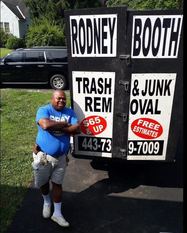 Rodney Booth stands next to his trash removal truck. He's smiling, wearing a blue shirt and shorts.