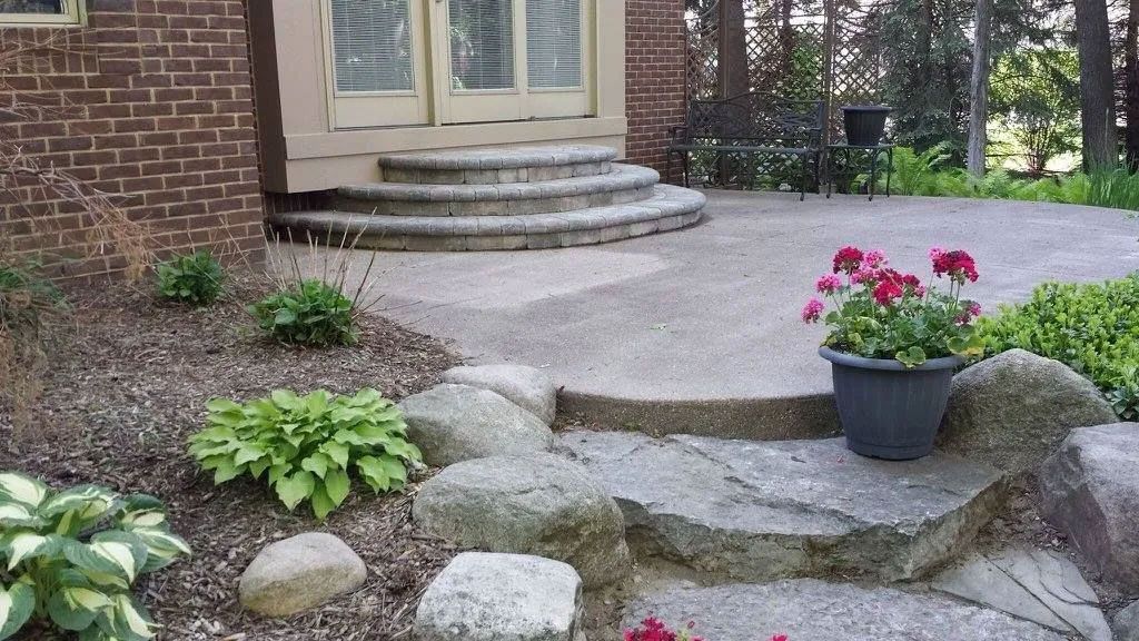 Stone steps lead to a brick house patio with a grey pot of pink flowers near large, natural landscape rocks.
