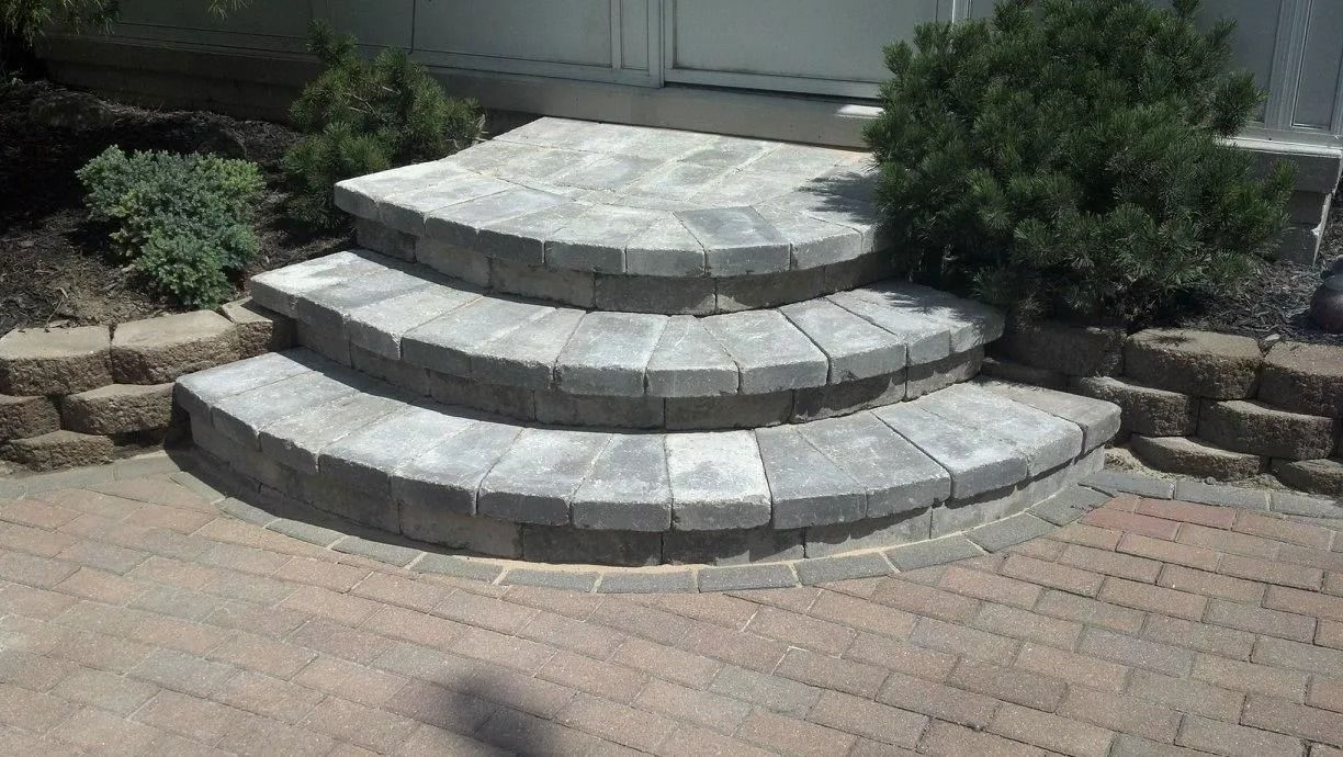 Three curved stone steps leading to a building entrance, flanked by landscaping stones and a small bush.