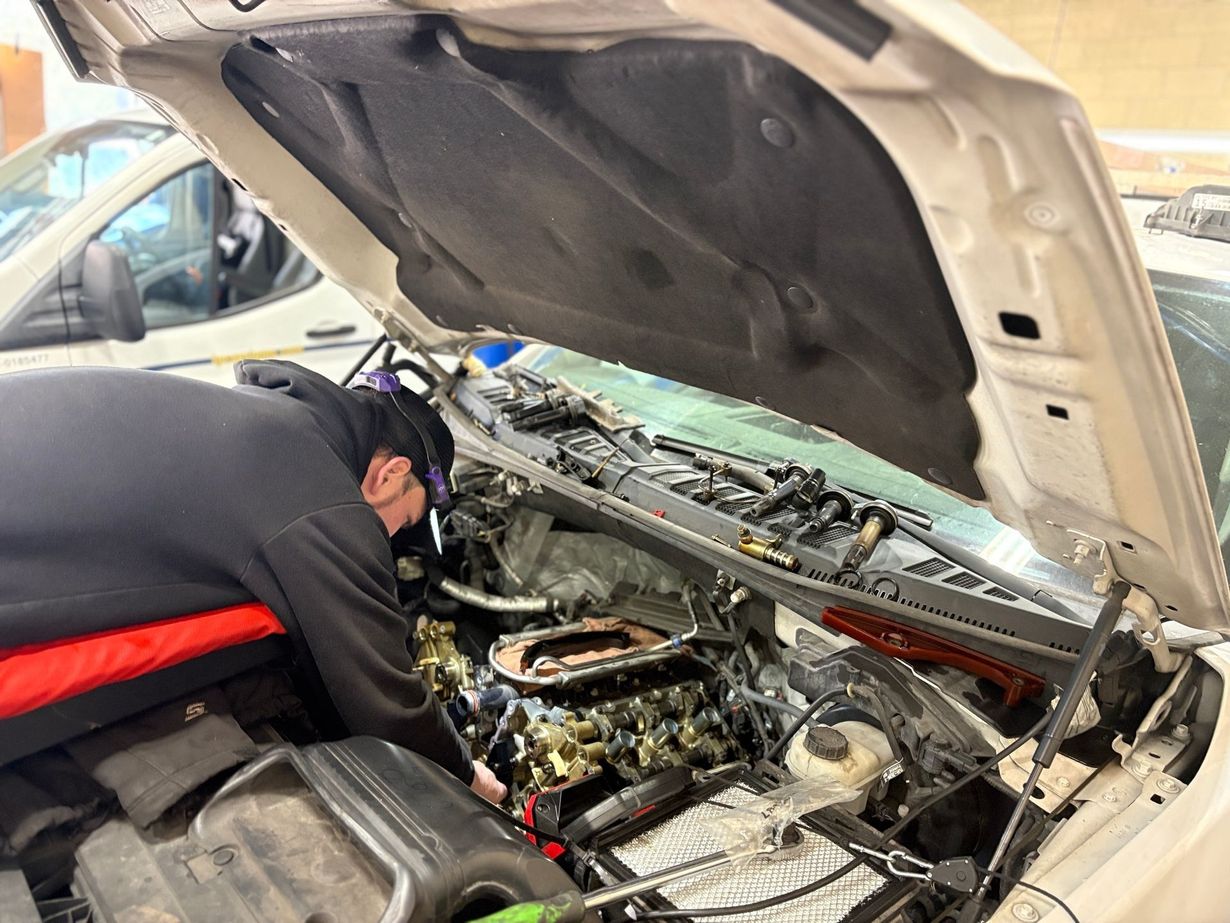 Mechanic working under an open car hood in a garage, tightening engine components with tools.