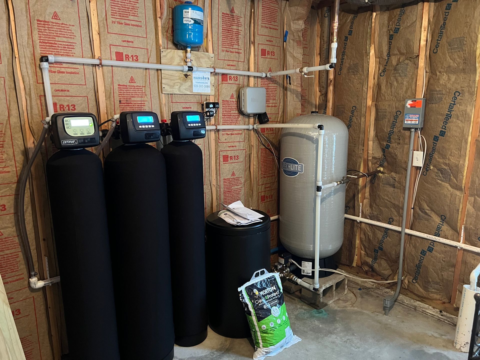 Water filtration system in a basement; three black tanks, one tall silver tank, blue pressure tank, exposed insulation.