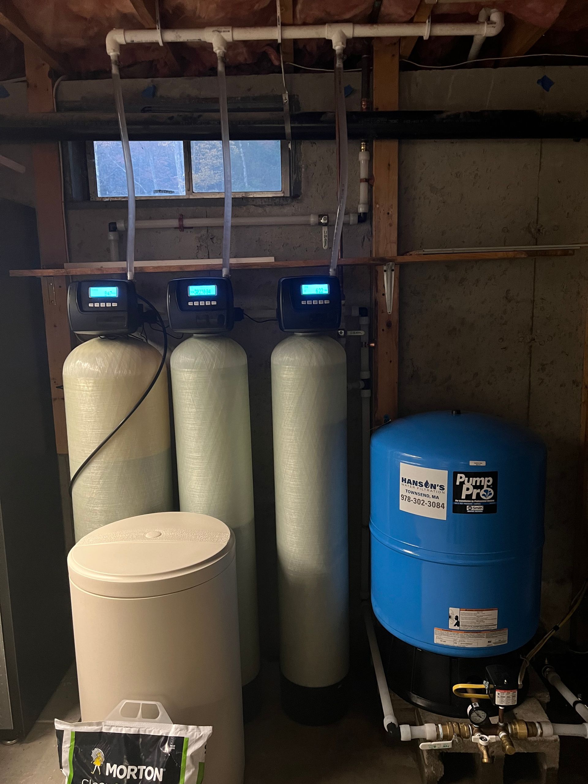 Water filtration system in a basement, with three tall white tanks, a blue pressure tank, and a salt container.