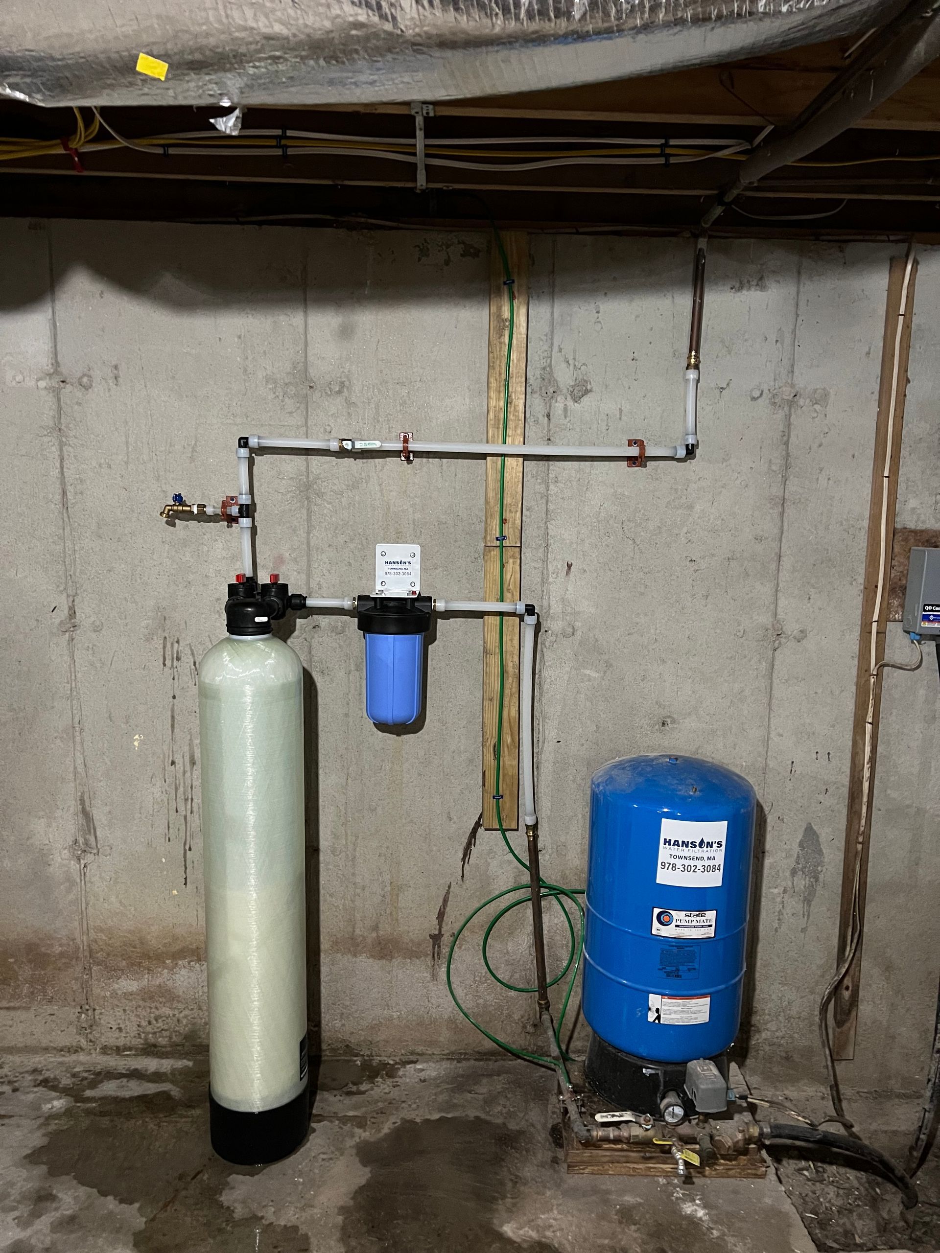 Water filtration system in a basement, including a tall white tank, blue filter, and blue pressure tank.