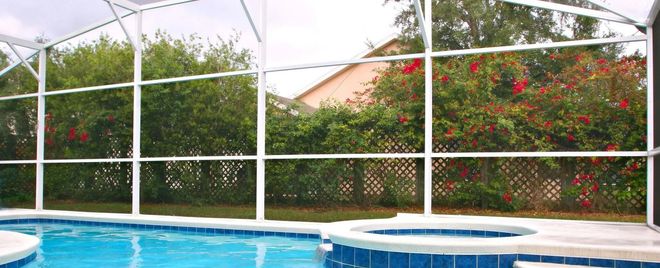 A screened-in pool area with a turquoise pool, spa, and lush green and red foliage.
