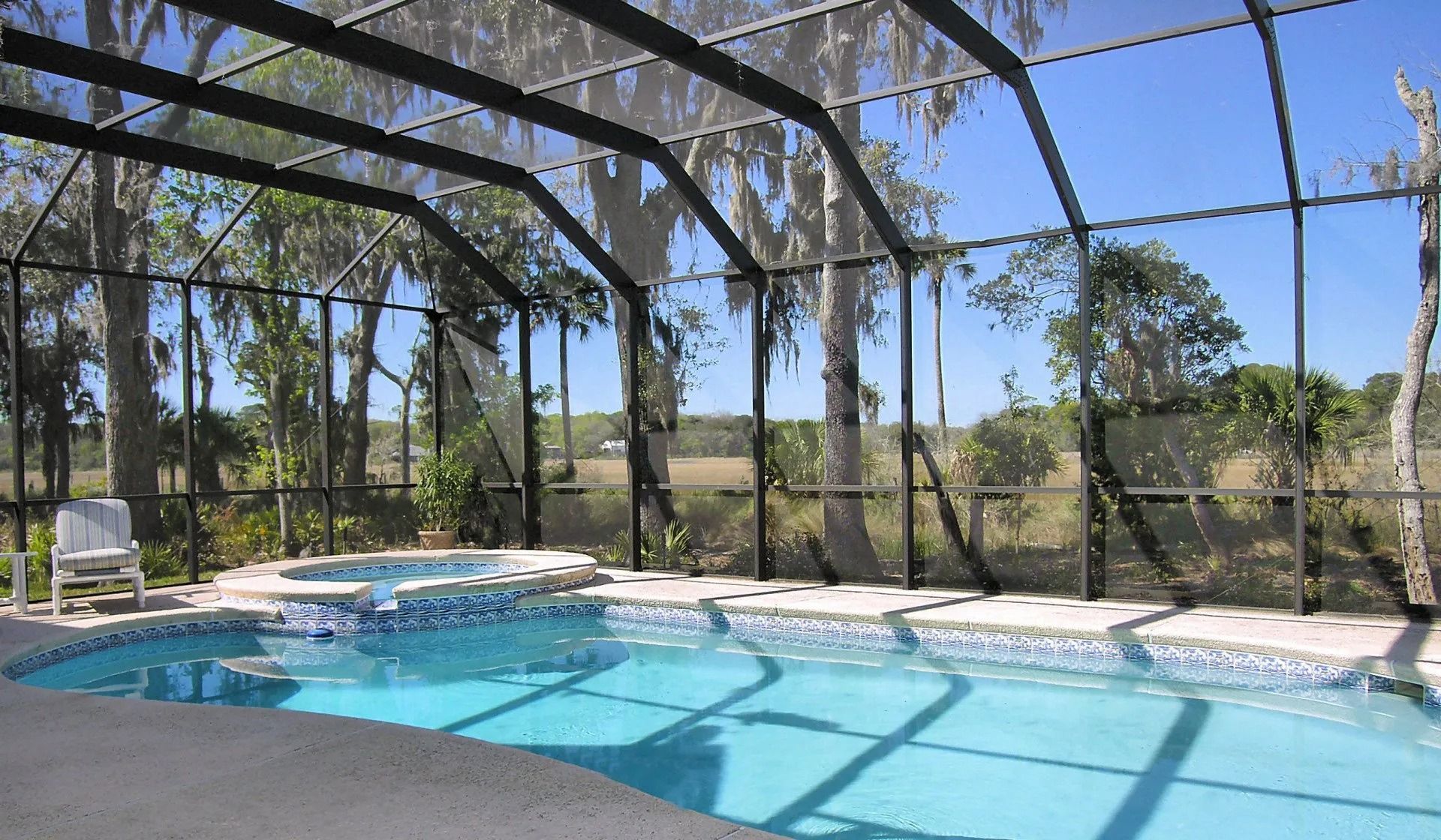 Pool and hot tub enclosed in a screened lanai with trees and fields in the background.