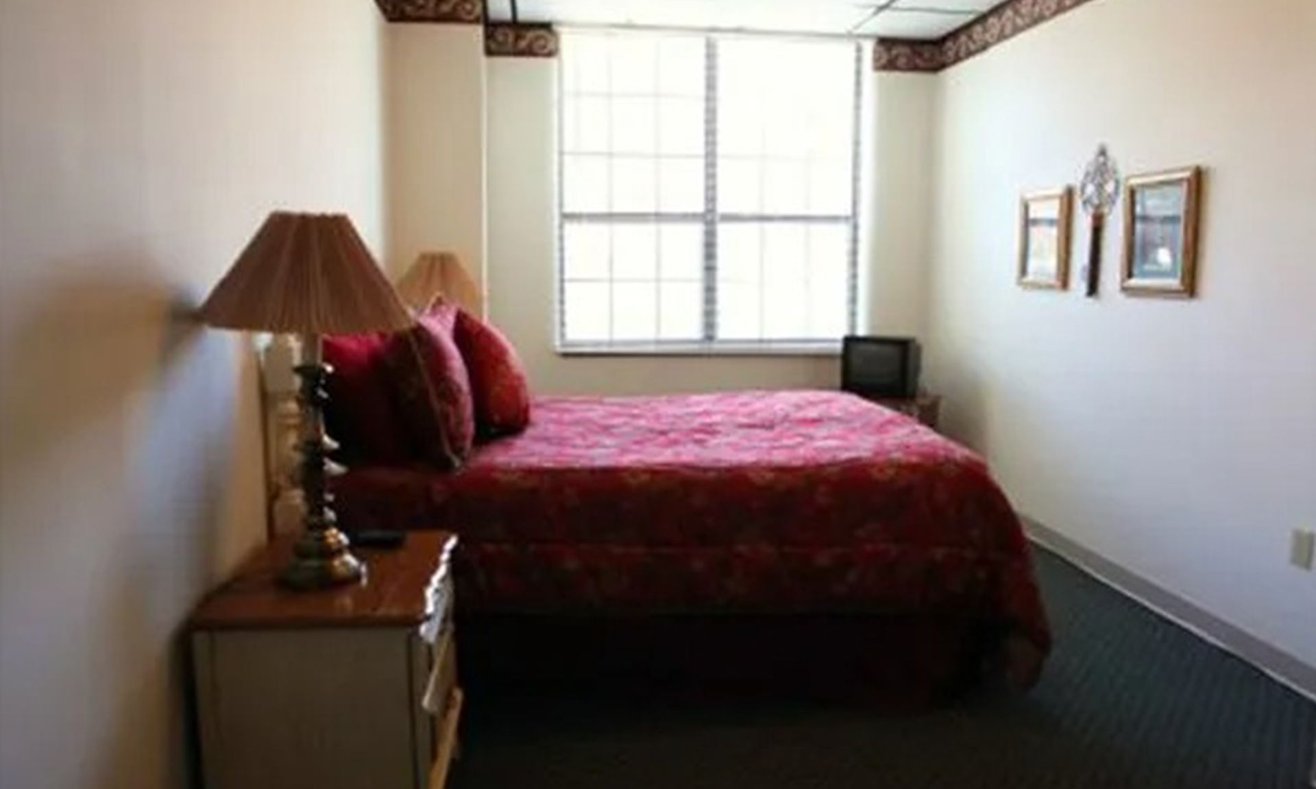 Bedroom with red patterned bedding, nightstand, lamp, window, small TV, and decorative art.