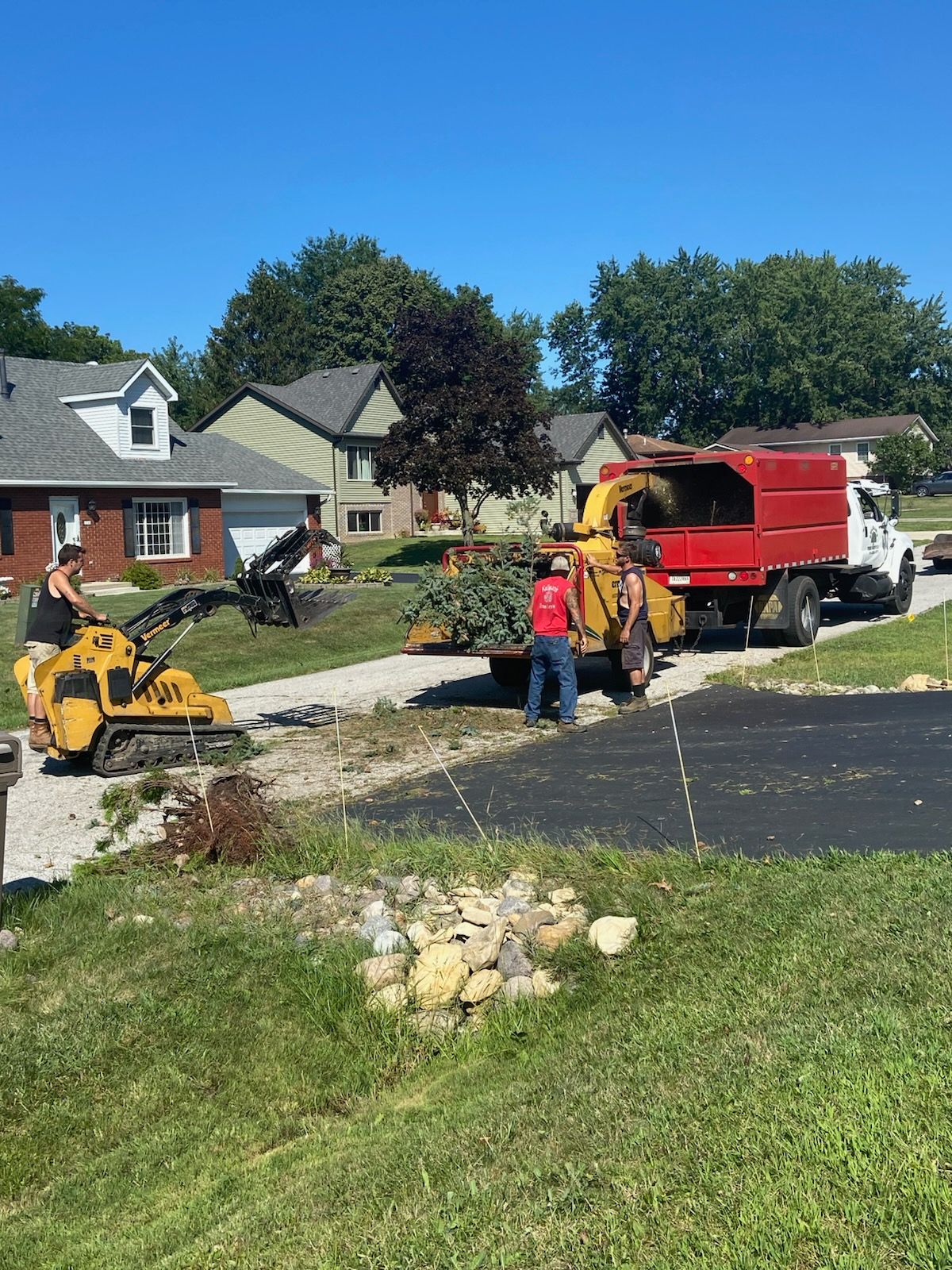 Men loading brush into a wood chipper on a residential driveway; sunny day.