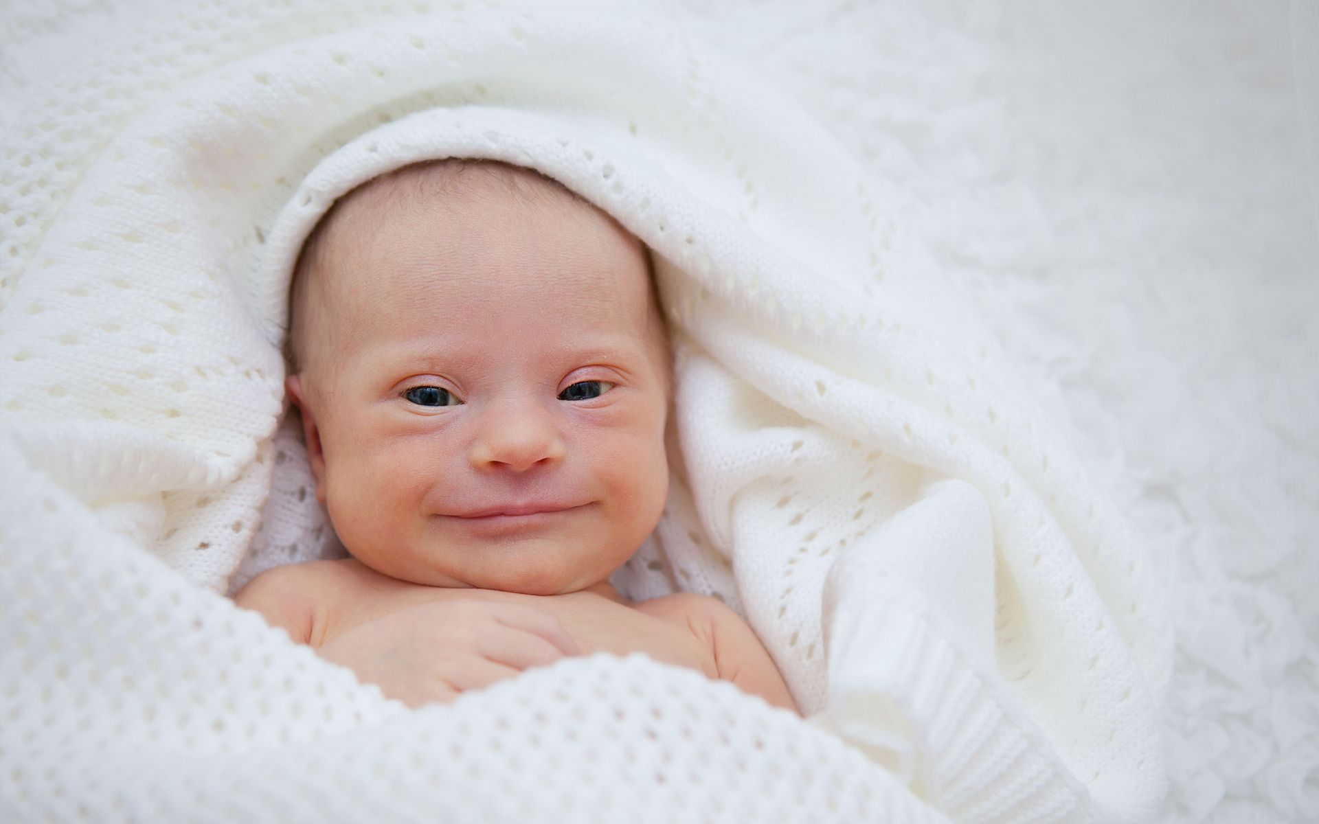 A baby with down syndrome is wrapped in a white blanket and smiling.