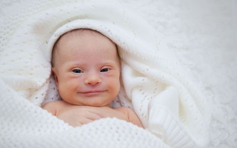 A baby with down syndrome is wrapped in a white blanket and smiling.